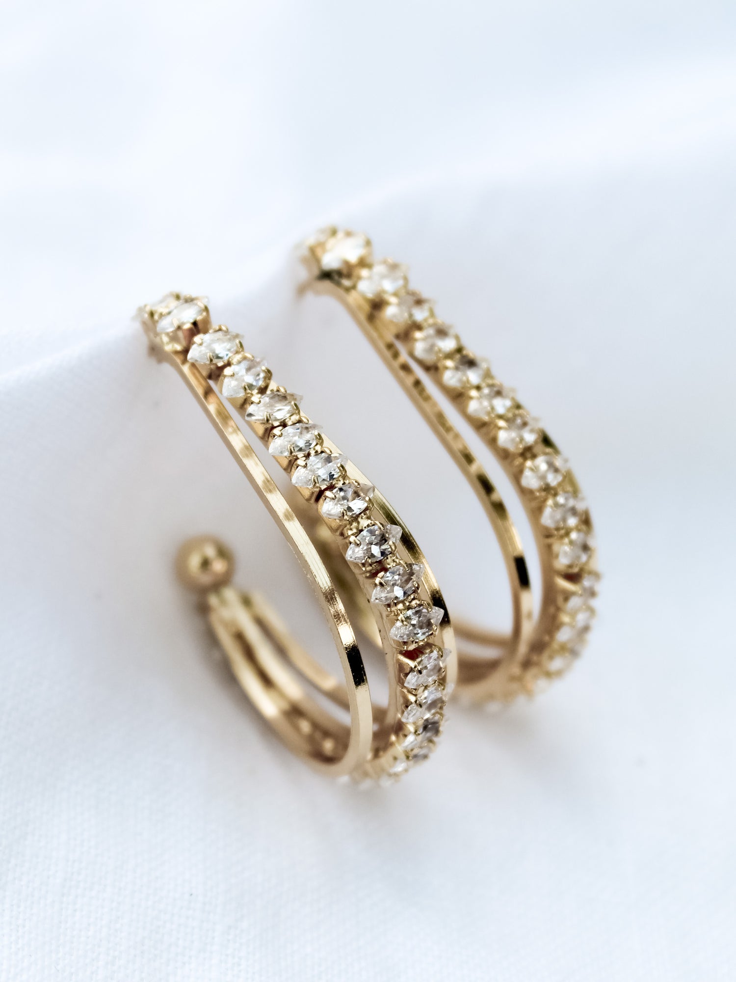 The Kenny Earring by Kinsey Designs Archives features gold-filled hoops adorned with two rows of sparkling cubic zirconia, displayed on a white fabric background.
