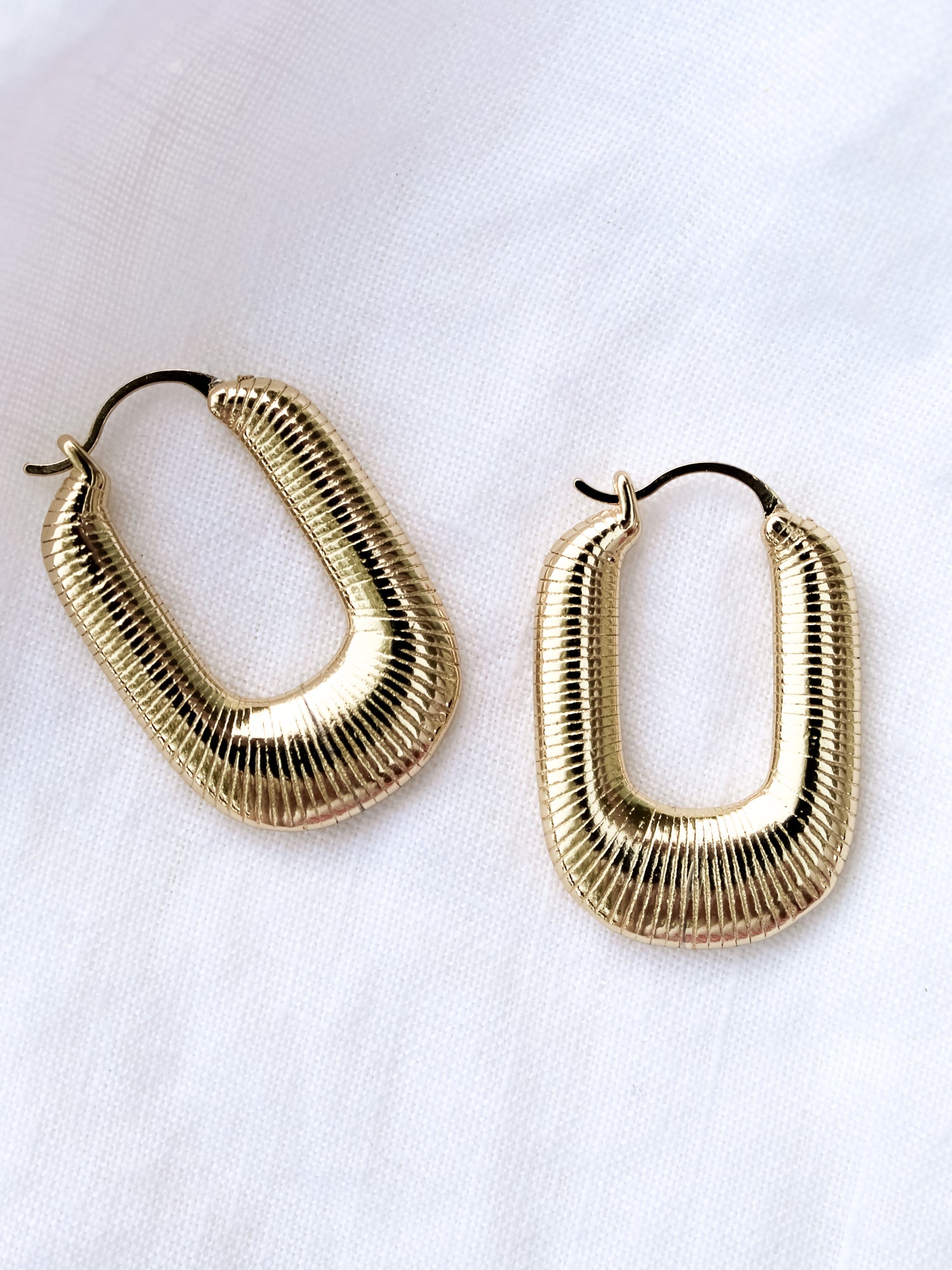 The Kepri Hoop by Kinsey Designs Archives features gold-plated, U-shaped hoops with a ribbed texture, measuring 1 inch long and displayed on a white fabric background.