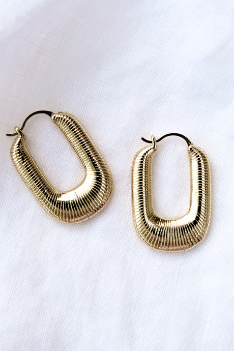 The Kepri Hoop by Kinsey Designs Archives features gold-plated, U-shaped hoops with a ribbed texture, measuring 1 inch long and displayed on a white fabric background.