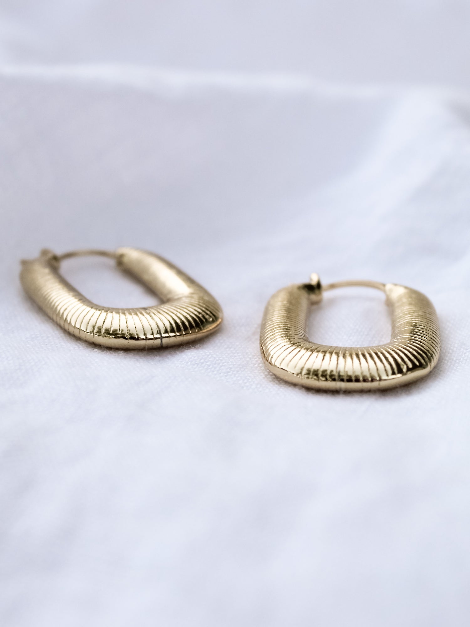 A pair of Kepri Hoop earrings by Kinsey Designs Archives, featuring gold plating and textured design, each measuring 1 inch, displayed on a soft white fabric background.