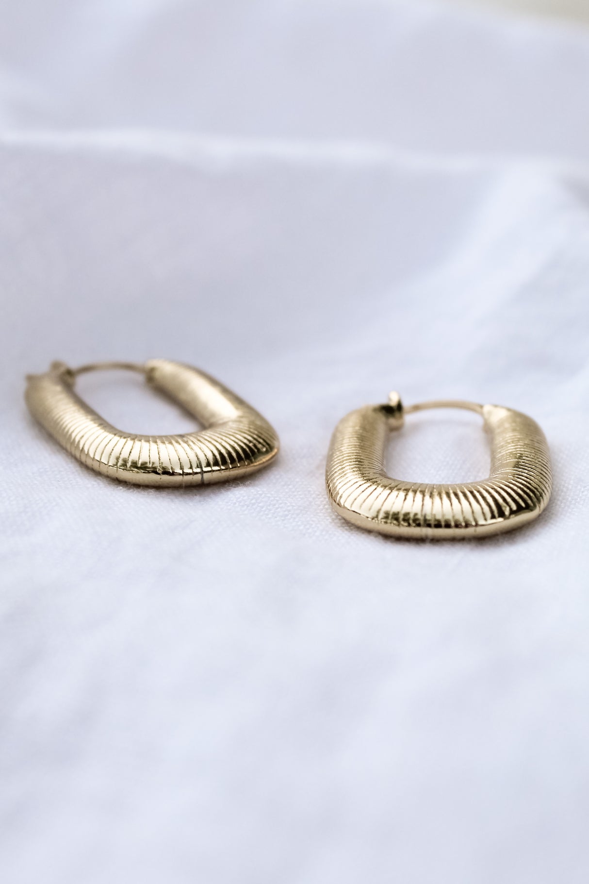 A pair of Kepri Hoop earrings by Kinsey Designs Archives, featuring gold plating and textured design, each measuring 1 inch, displayed on a soft white fabric background.