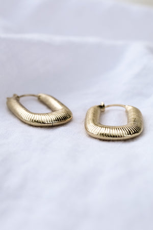 A pair of Kepri Hoop earrings by Kinsey Designs Archives, featuring gold plating and textured design, each measuring 1 inch, displayed on a soft white fabric background.