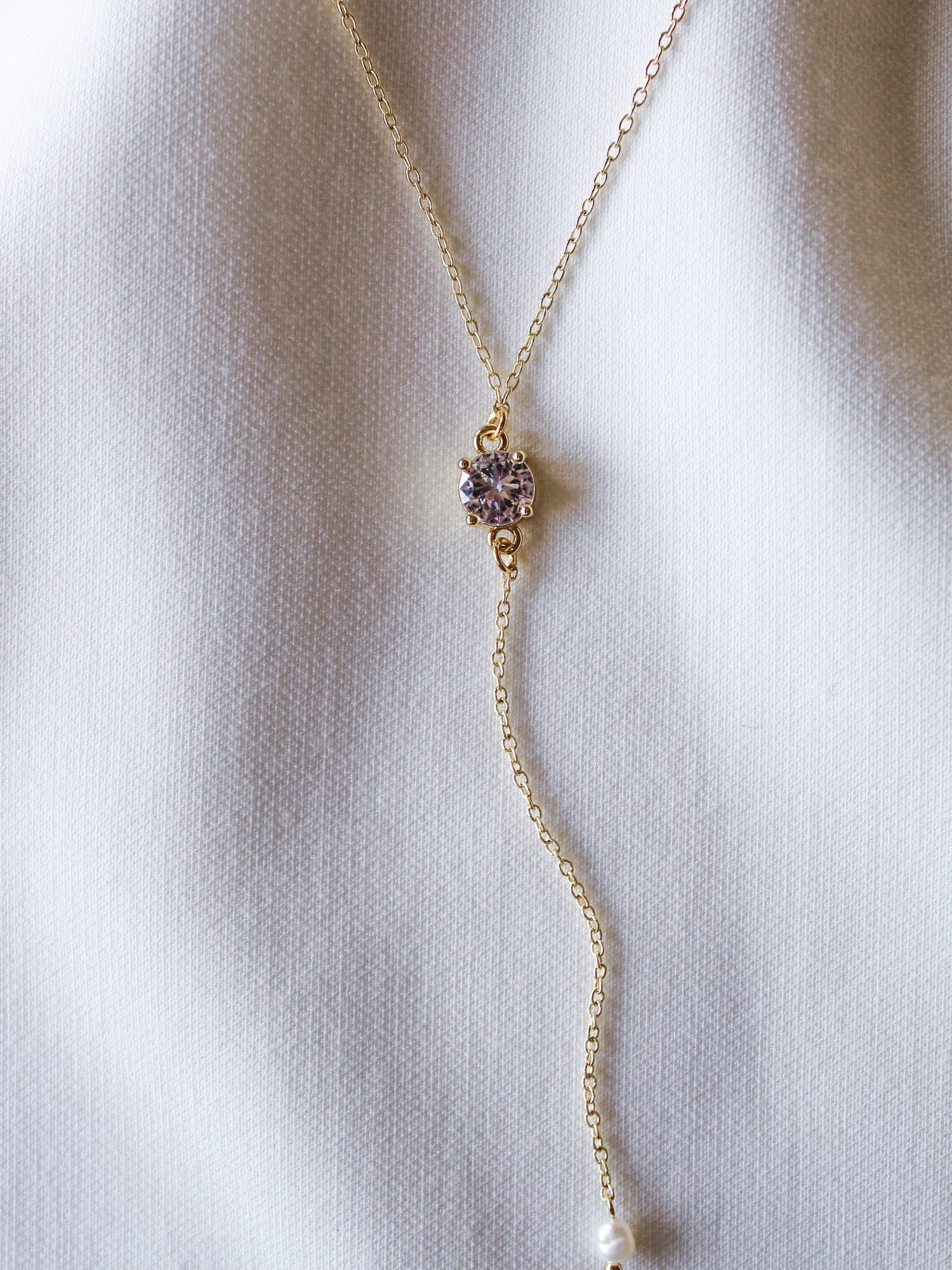 The Logan Lariat by Kinsey Designs Archives is a gold filled necklace with a small purple cubic zirconia and pearl dangle, showcased on a white fabric background.