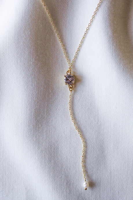 The Logan Lariat by Kinsey Designs Archives is a gold filled necklace with a small purple cubic zirconia and pearl dangle, showcased on a white fabric background.