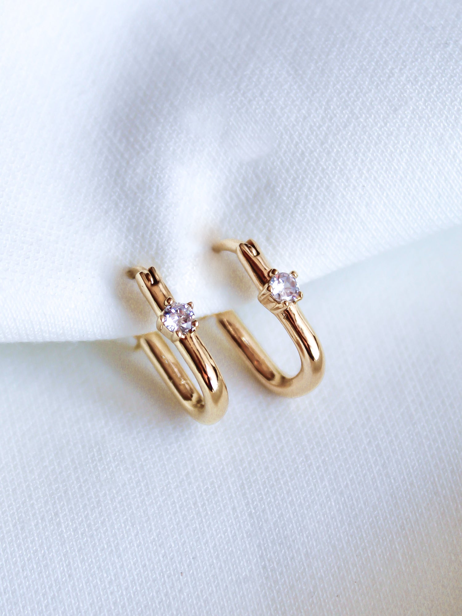 The Lou Hoop by Kinsey Designs Archives features 14k gold plating and small clear cubic zirconia gemstones, elegantly displayed on white fabric.