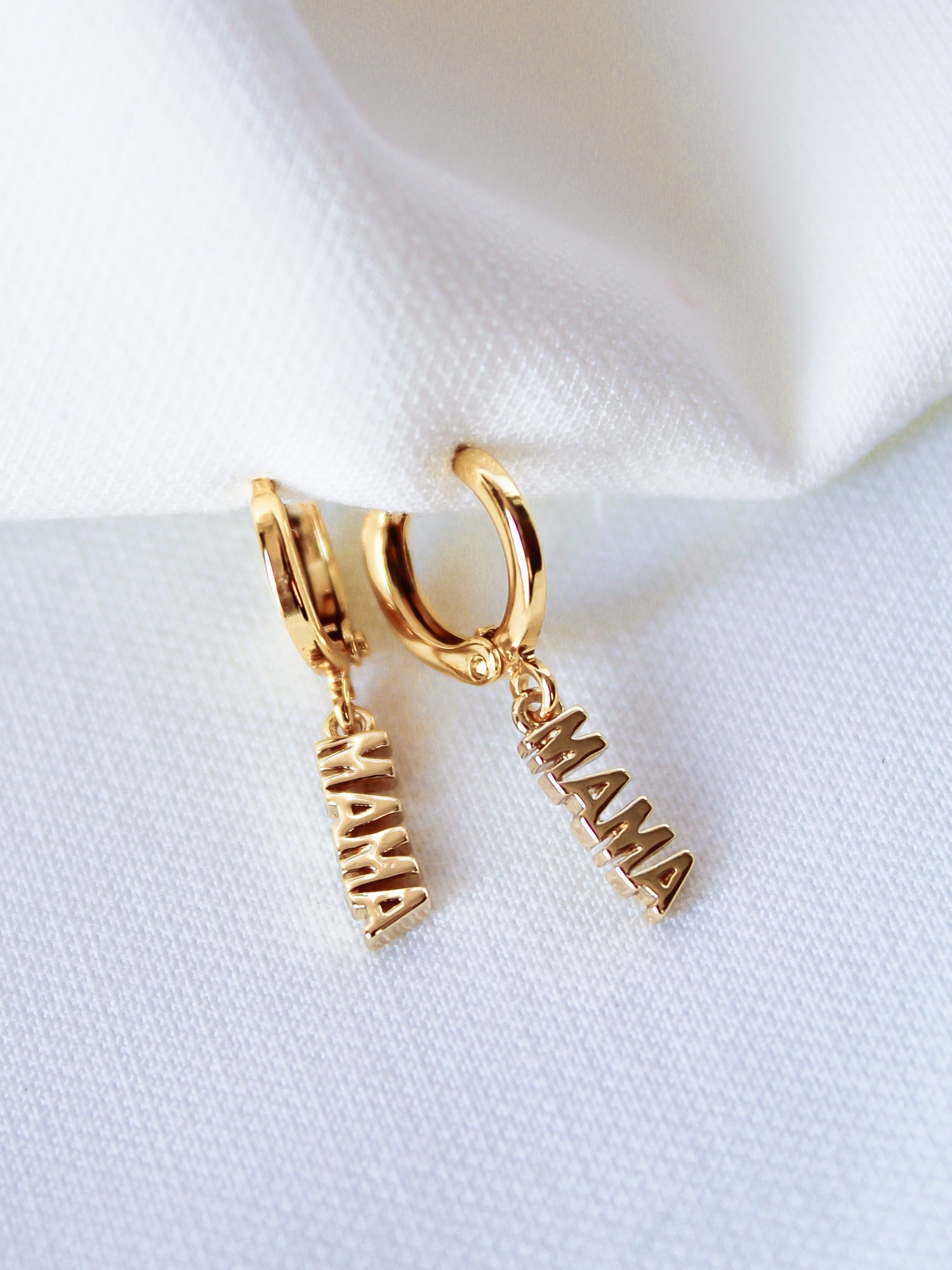 The MAMA Huggies by Kinsey Designs Archives are 1-inch gold filled hoop earrings with "MAMA" letter charms, shown on a white fabric background.