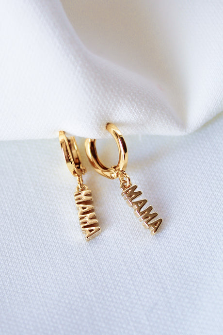 The MAMA Huggies by Kinsey Designs Archives are 1-inch gold filled hoop earrings with "MAMA" letter charms, shown on a white fabric background.
