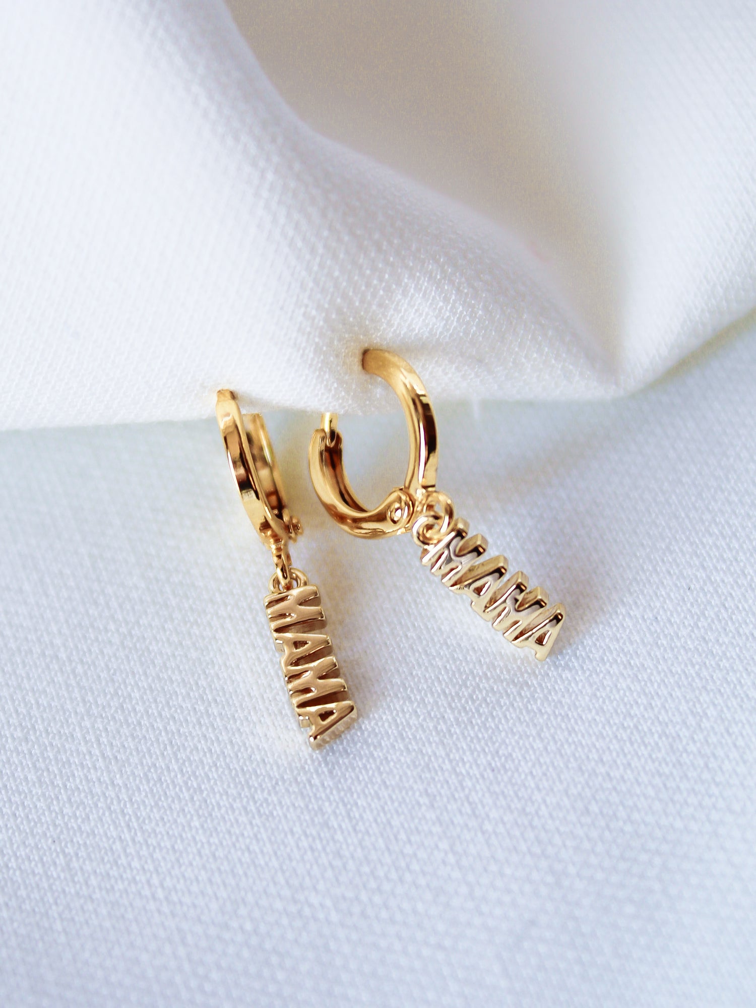 The MAMA Huggies by Kinsey Designs Archives are 1-inch gold filled earrings featuring "MAMA" letter charms, elegantly displayed on white fabric.