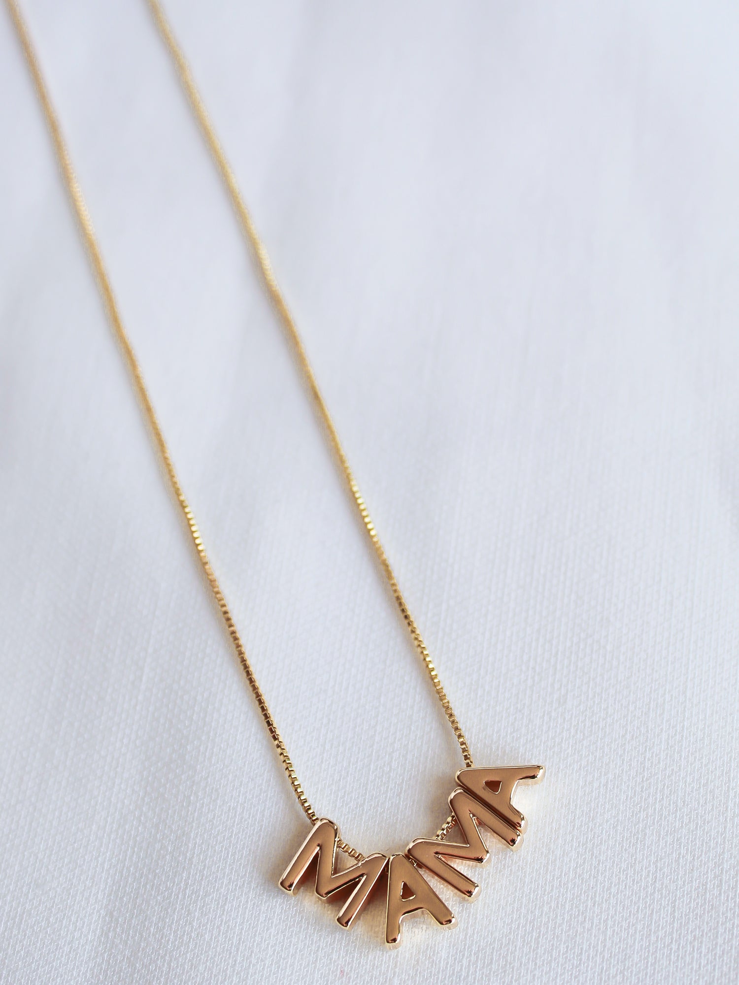 The MAMA Slide Necklace by Kinsey Designs Archives features gold letter charms spelling "MAMA" on a white fabric background, with a 14k gold filled 18" chain and an adjustable Italian closure.