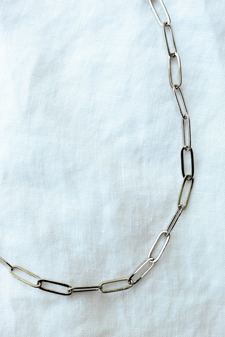The Maeve Silver by Kinsey Designs Archives features a thin, silver chain with elongated oval links and a secure lobster clasp, displayed against a white fabric background.
