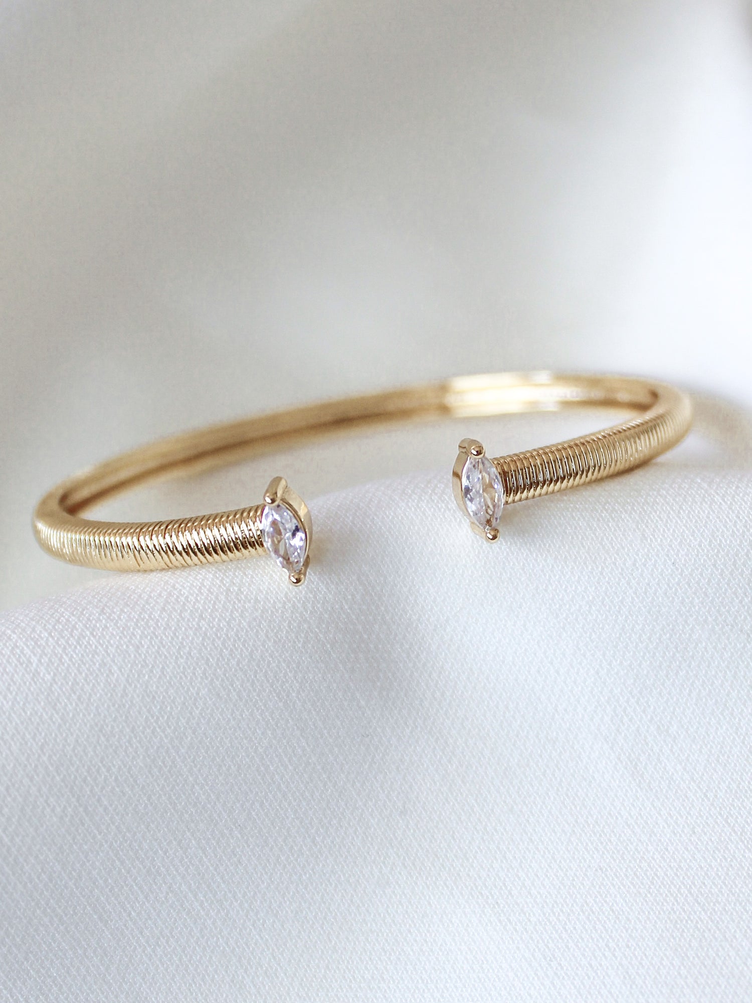 The Mara Cuff by Kinsey Designs is a gold plated adjustable bracelet, showcasing two clear cubic zirconia gemstones on each end, displayed on a white fabric background.