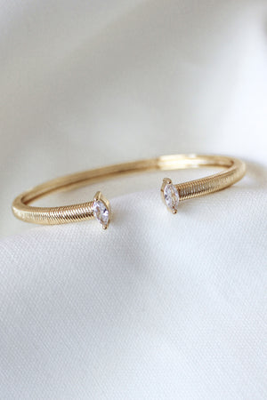 The Mara Cuff by Kinsey Designs is a gold plated adjustable bracelet, showcasing two clear cubic zirconia gemstones on each end, displayed on a white fabric background.