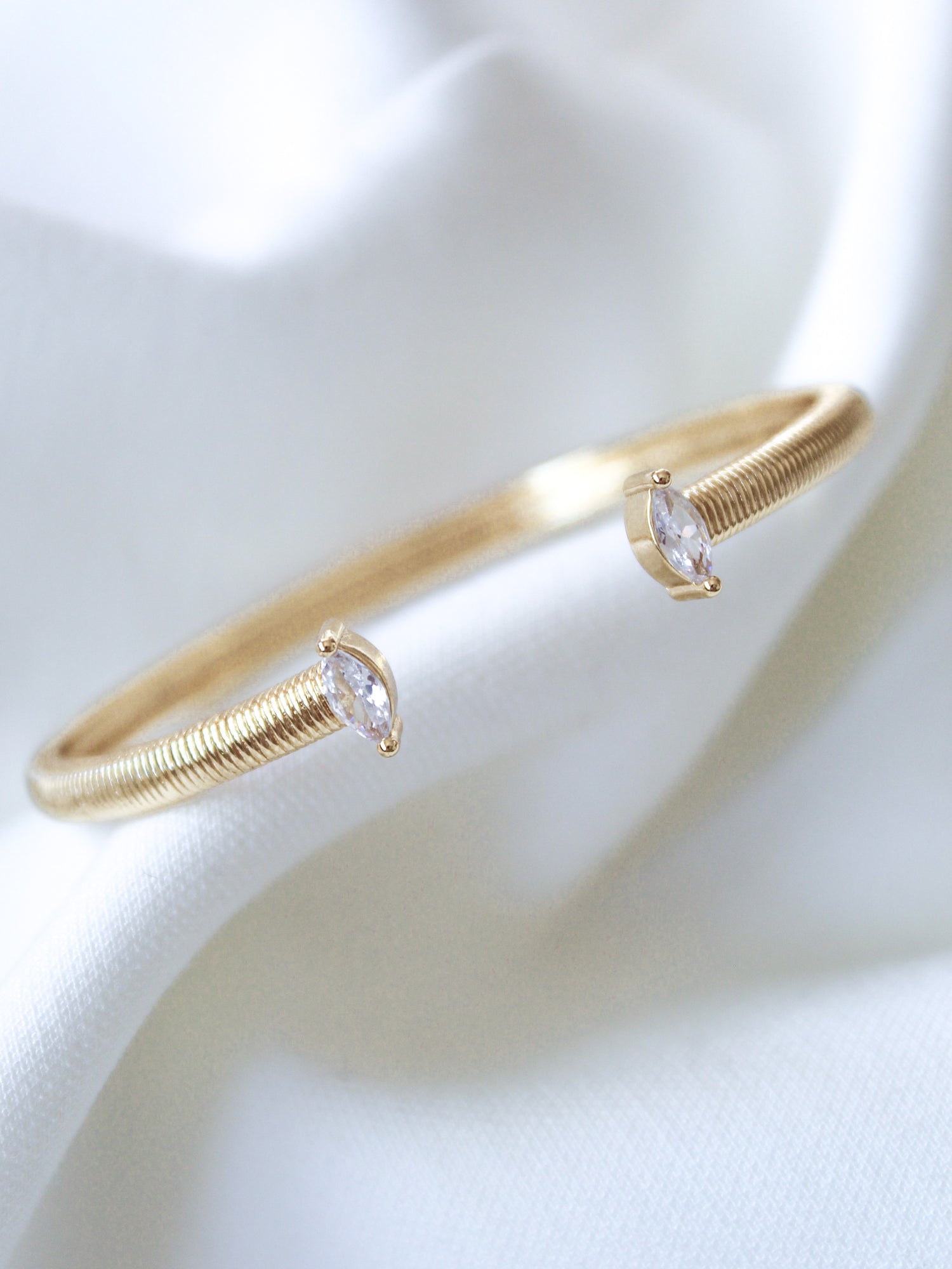 The Mara Cuff by Kinsey Designs is a gold-plated bracelet featuring two marquise-cut cubic zirconia stones, elegantly displayed on soft white fabric and adjustable for a perfect fit.