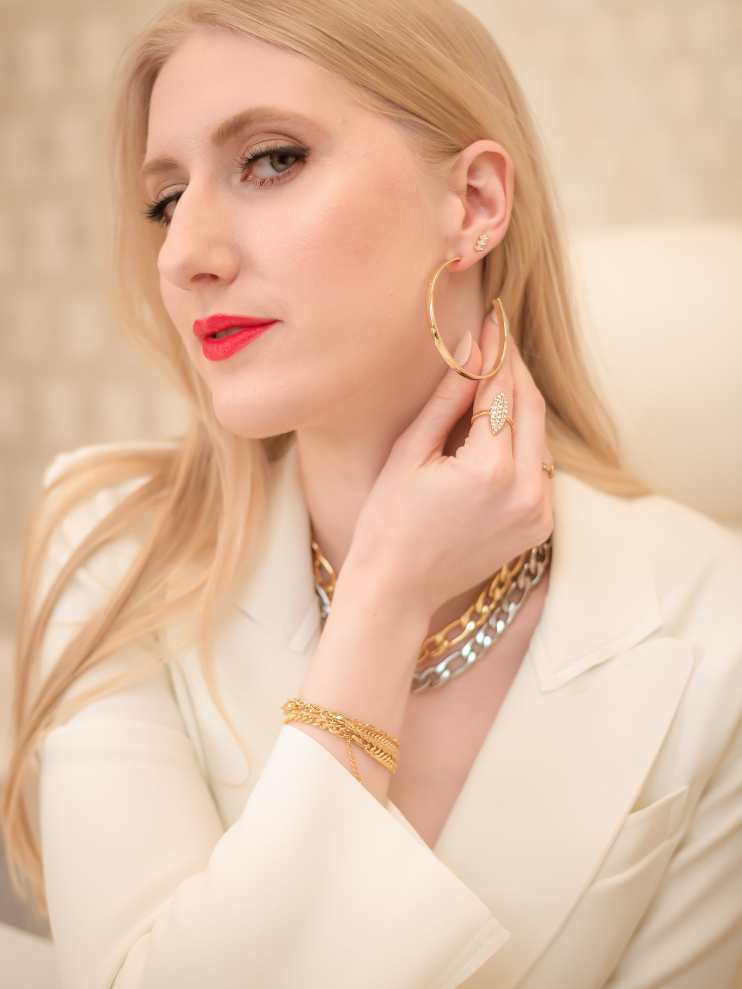 A woman with blonde hair in a white blazer touches her Kinsey Designs Archives Molly Hoop gold plated earrings, styled with gold jewelry and red lipstick.