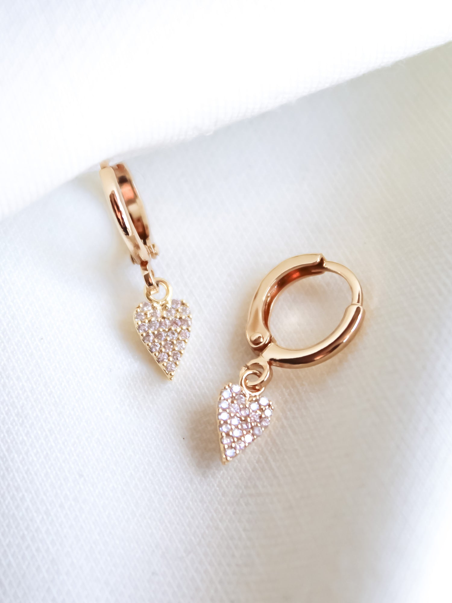 The Naomi Huggies by Kinsey Designs Archives are gold hoop earrings featuring small, sparkling heart-shaped cubic zirconia pendants with a 14k gold plated finish, shown against a white fabric background.
