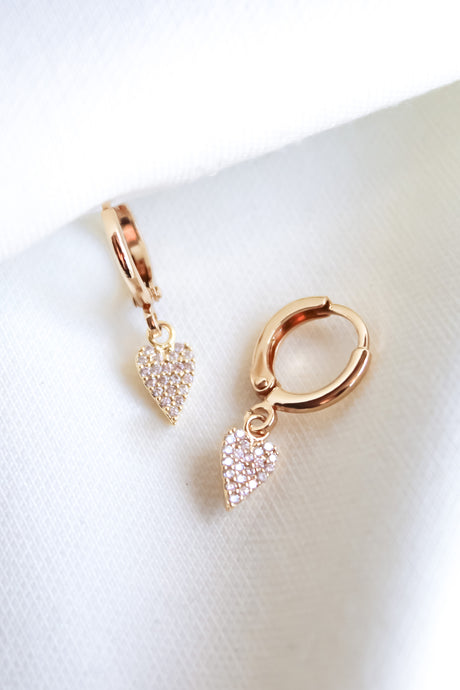 The Naomi Huggies by Kinsey Designs Archives are gold hoop earrings featuring small, sparkling heart-shaped cubic zirconia pendants with a 14k gold plated finish, shown against a white fabric background.