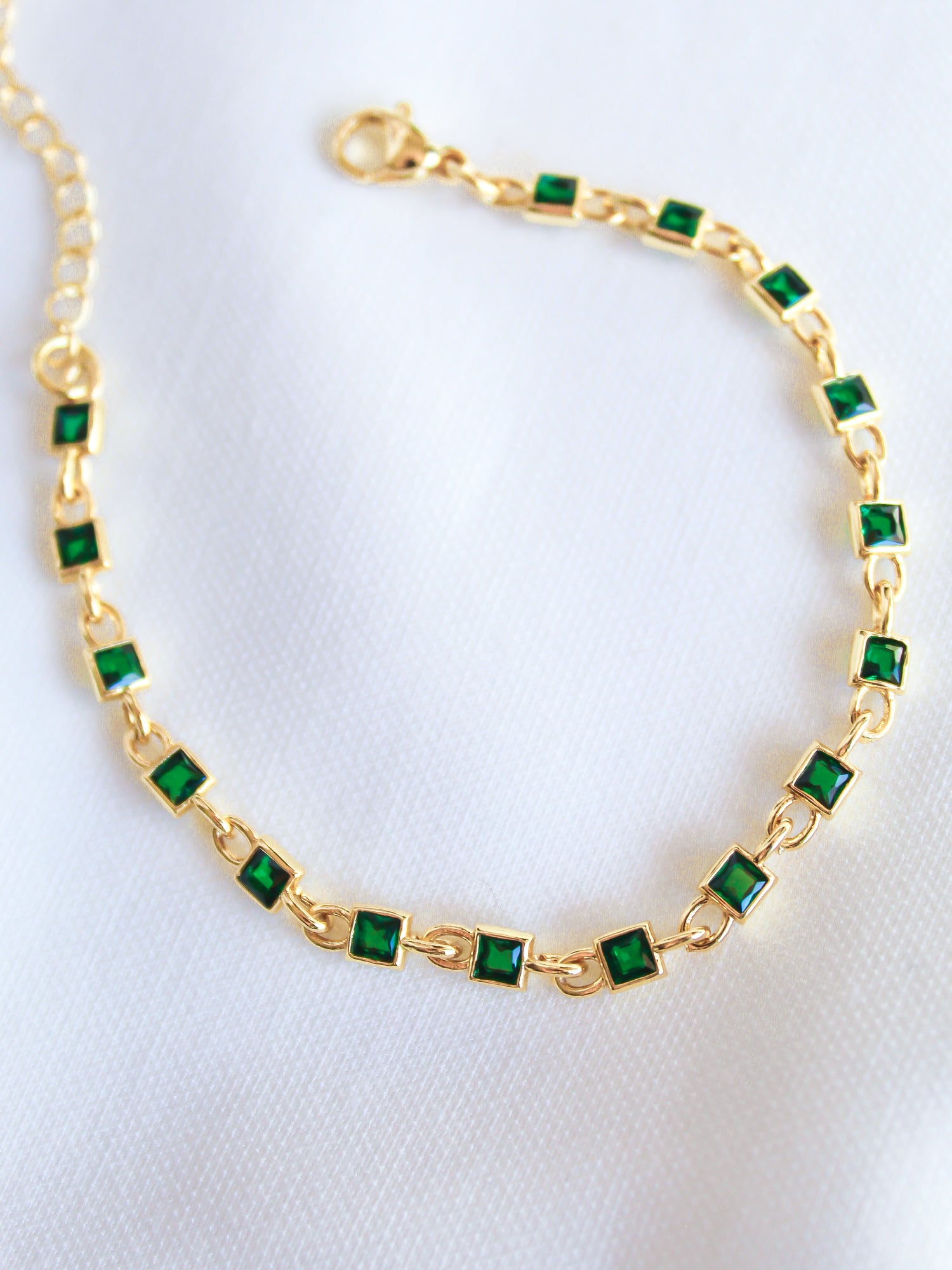 The Paddy Bracelet by Kinsey Designs Archives features square green gemstones and an adjustable gold filled chain, shown on a white fabric background.