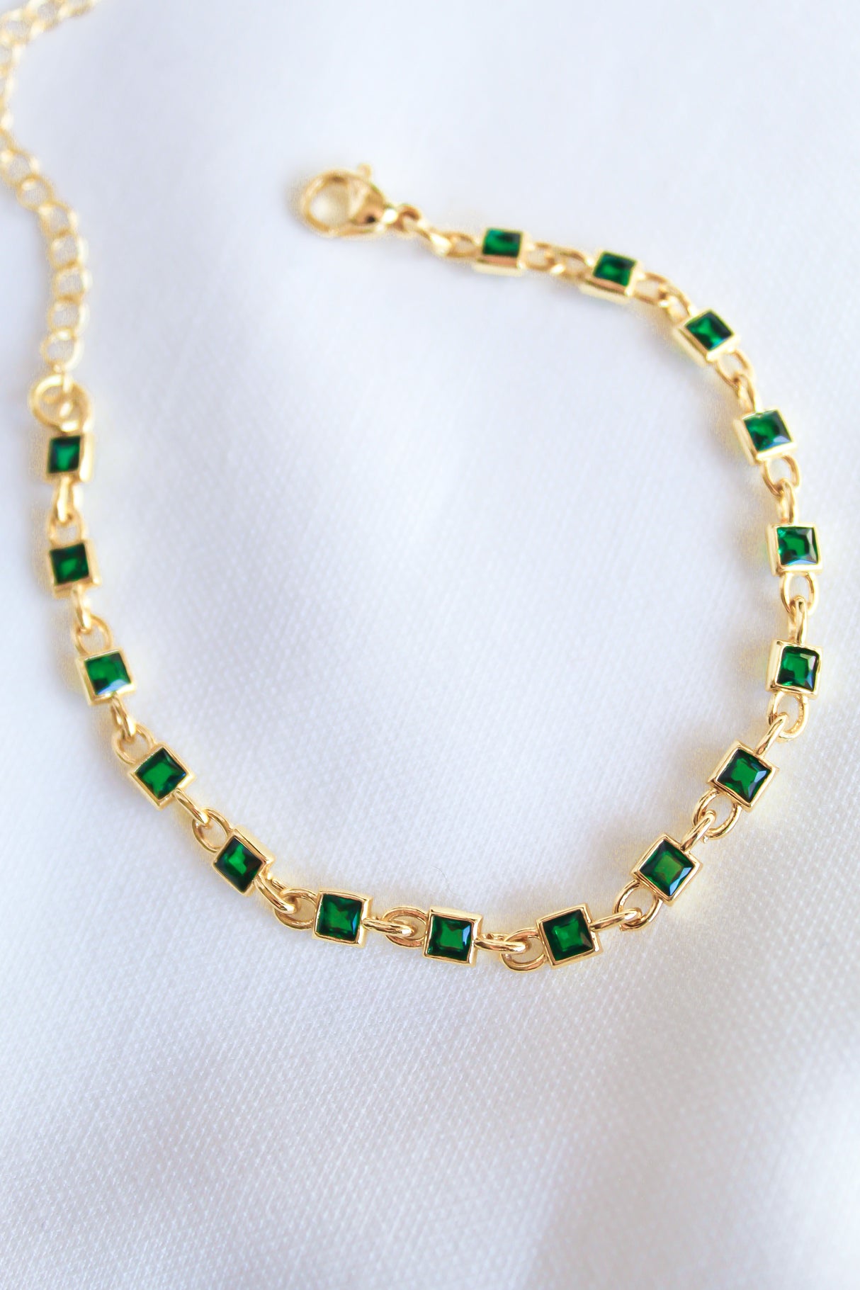 The Paddy Bracelet by Kinsey Designs Archives features square green gemstones and an adjustable gold filled chain, shown on a white fabric background.