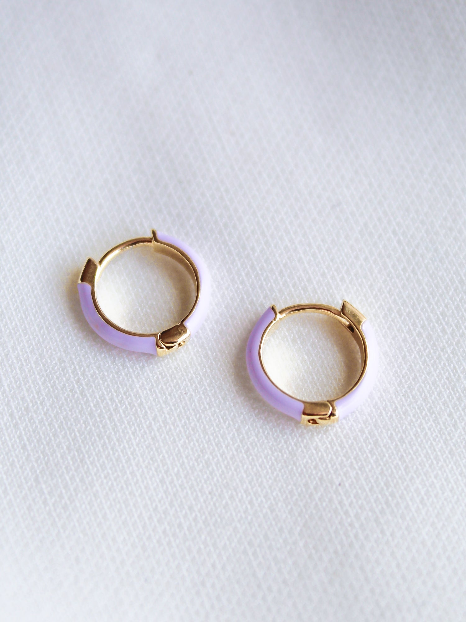 The Pear Huggie - Lavender by Kinsey Designs Archives features gold plated hoop earrings with light purple enamel accents and a 0.5" diameter, shown on a white textured fabric background.