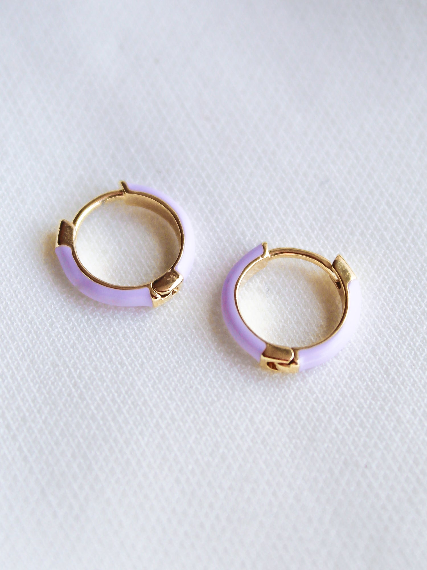 The Pear Huggie - Lavender by Kinsey Designs Archives features two small gold plated hoops with light purple enamel accents, each .5" in diameter, displayed on a white textured surface.