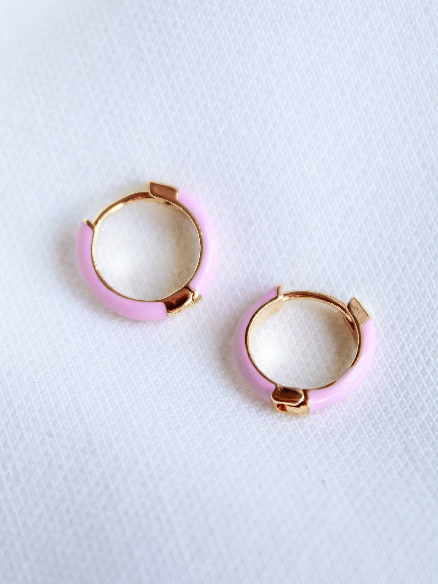 The Kinsey Designs Archives Pear Huggie - Pink features two small gold plated hoop earrings with pastel pink enamel and a .5" diameter, displayed on a white textured fabric background.
