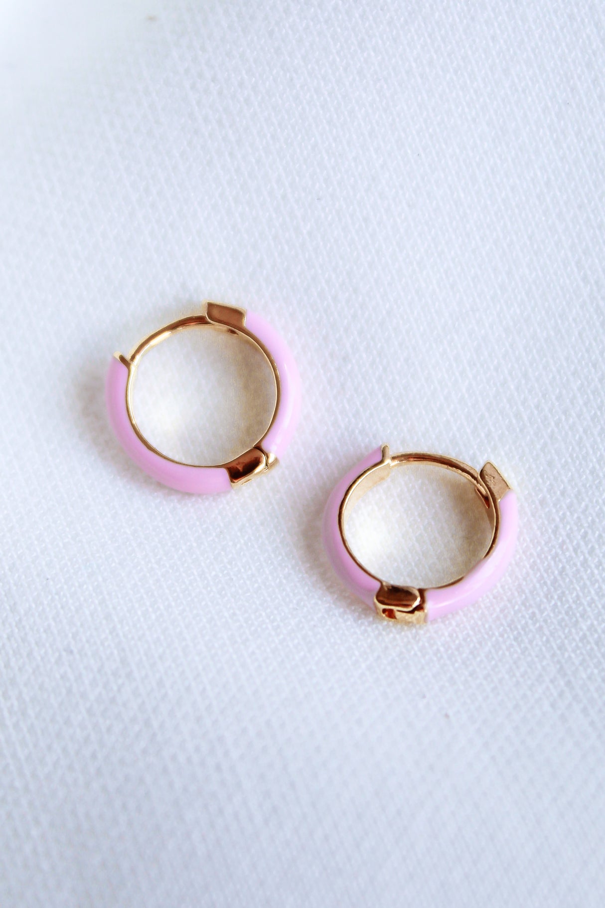 The Kinsey Designs Archives Pear Huggie - Pink features two small gold plated hoop earrings with pastel pink enamel and a .5" diameter, displayed on a white textured fabric background.