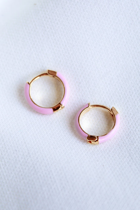 The Kinsey Designs Archives Pear Huggie - Pink features two small gold plated hoop earrings with pastel pink enamel and a .5" diameter, displayed on a white textured fabric background.