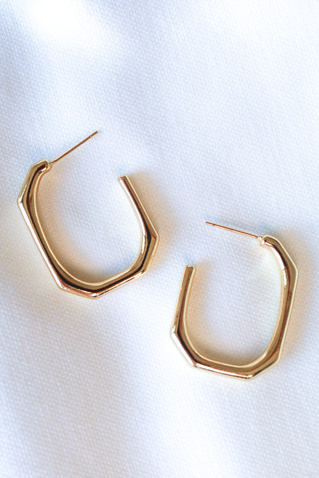 The Polly Hoop by Kinsey Designs Archives is a pair of gold plated hexagonal hoop earrings, measuring 1" wide and 1.25" long, shown on a white fabric background.