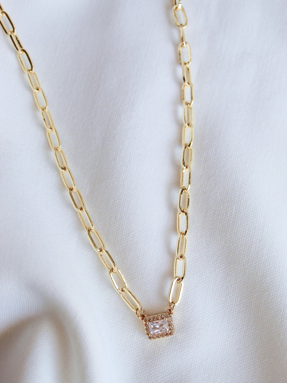 The Posh Necklace by Kinsey Designs features a gold-filled rectangular pendant, elegantly displayed on white fabric—a perfect statement piece for any occasion.
