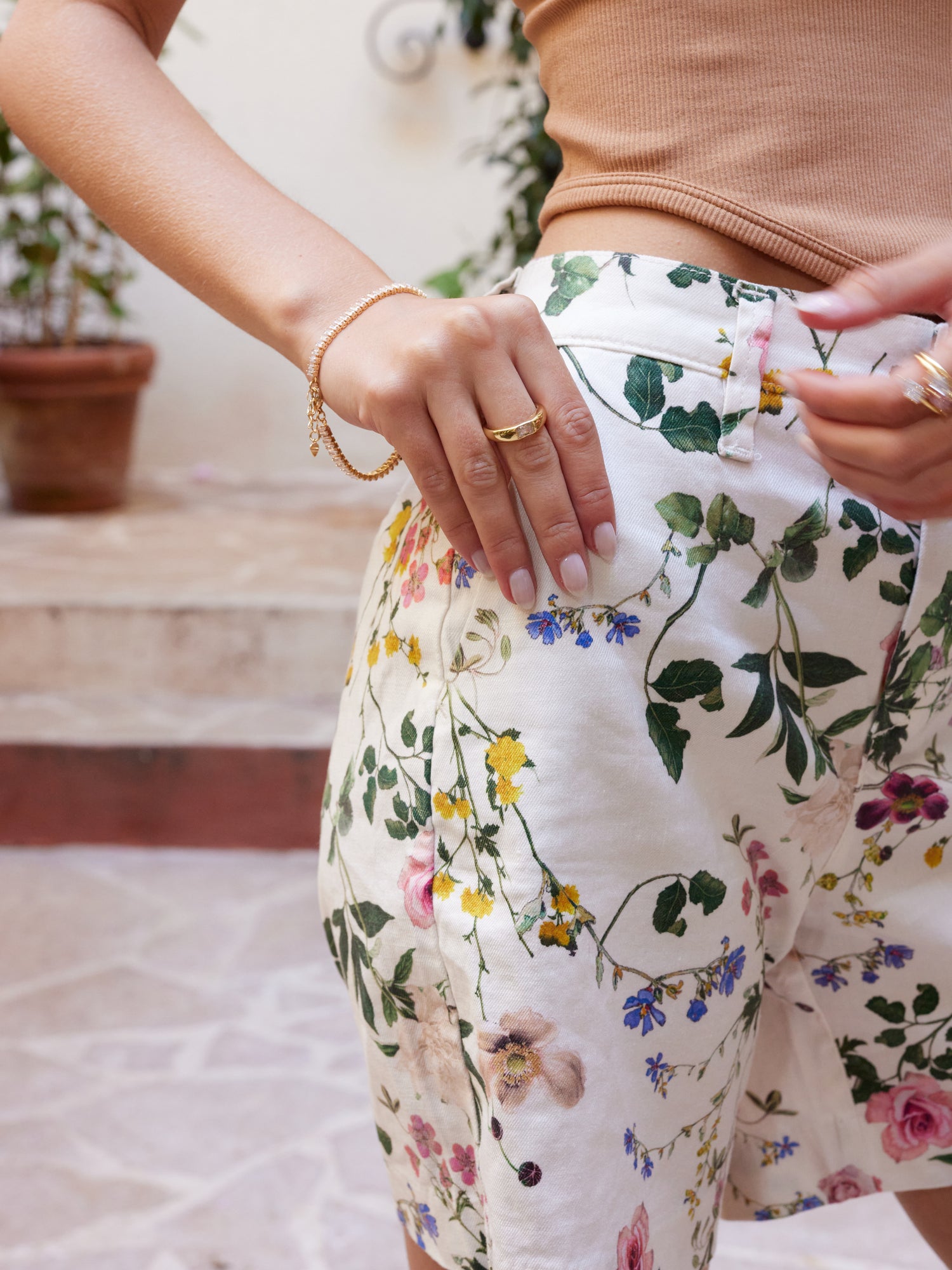 A woman wearing floral shorts and a tan top stands outdoors, showcasing the Preston Ring by Kinsey Designs Archives—a gold plated piece adorned with sparkling cubic zirconia.