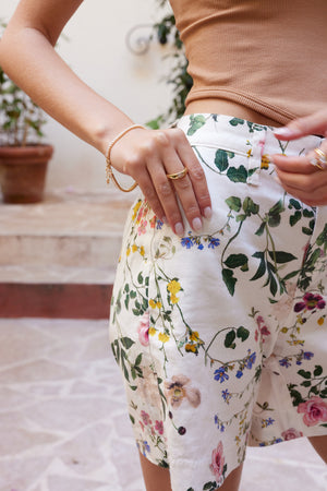 A woman wearing floral shorts and a tan top stands outdoors, showcasing the Preston Ring by Kinsey Designs Archives—a gold plated piece adorned with sparkling cubic zirconia.