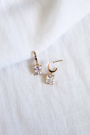 The Kinsey Designs Prism Huggies, featuring two gold huggie earrings adorned with princess-cut CZ gemstones, are displayed on white fabric.