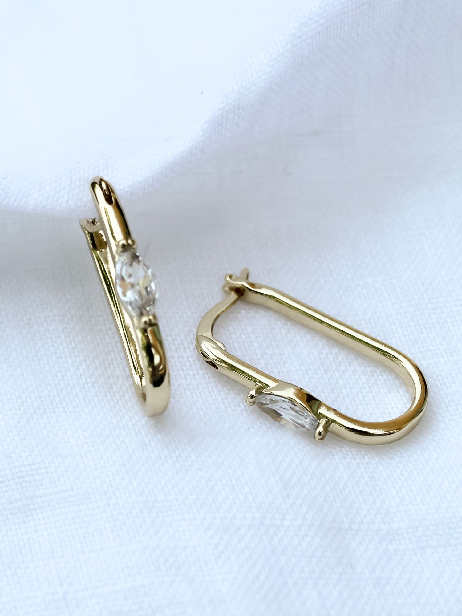 The Ragan Huggie earrings by Kinsey Designs Archives feature gold-filled hoops, 1" long, with sparkling cubic zirconia gemstones, shown on a white fabric background.