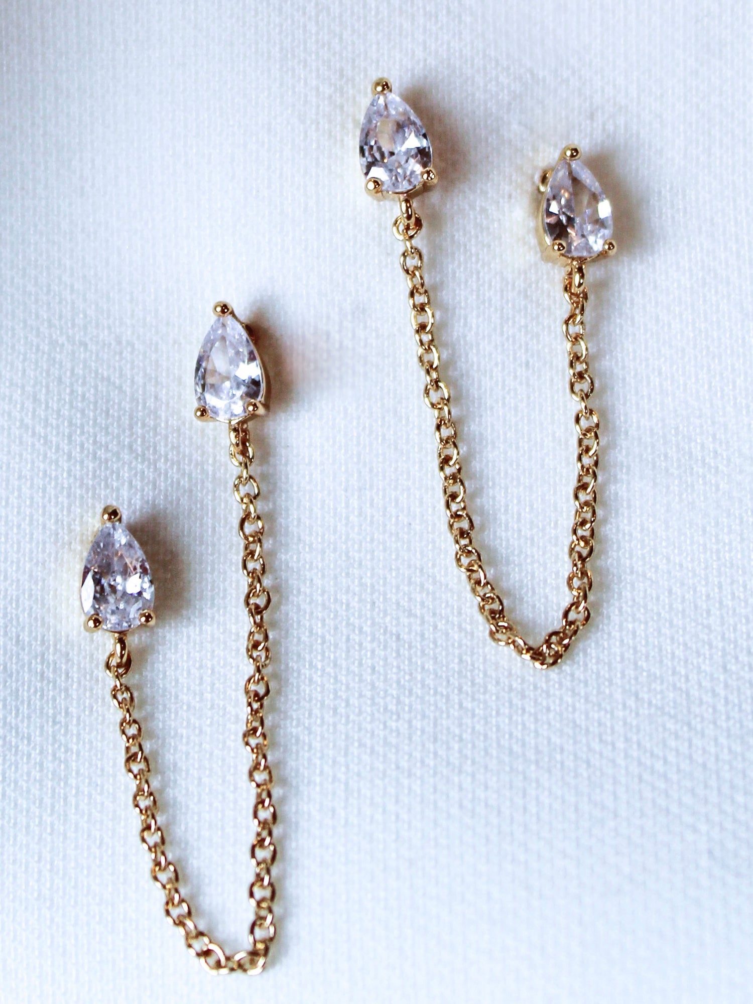 The Rayna Post earrings by Kinsey Designs Archives, featuring gold-plated chains and teardrop-shaped cubic zirconia, are displayed on a light fabric background.