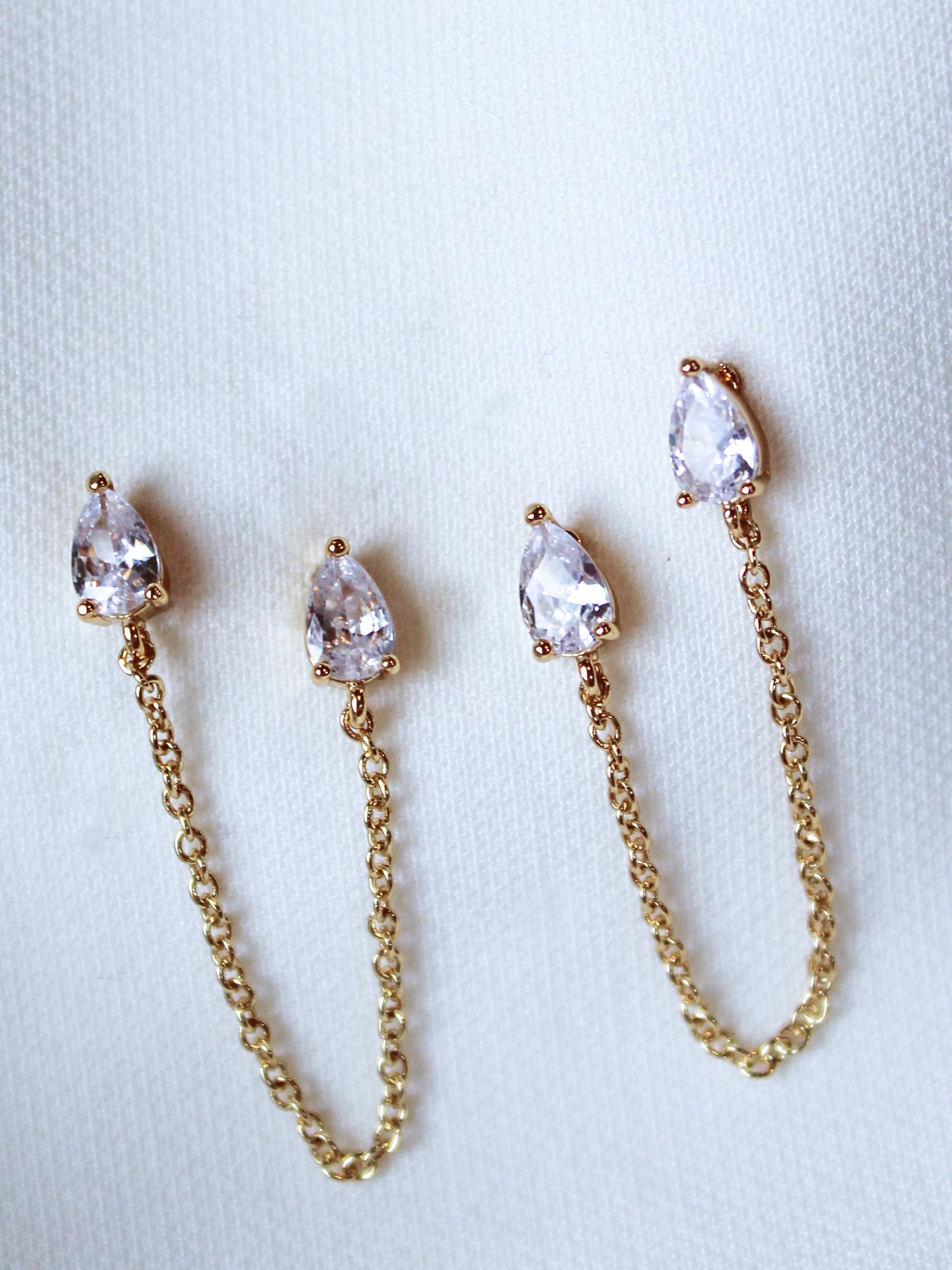 The Rayna Post by Kinsey Designs Archives are gold plated chain earrings with teardrop-shaped cubic zirconia, elegantly shown on a white fabric background.