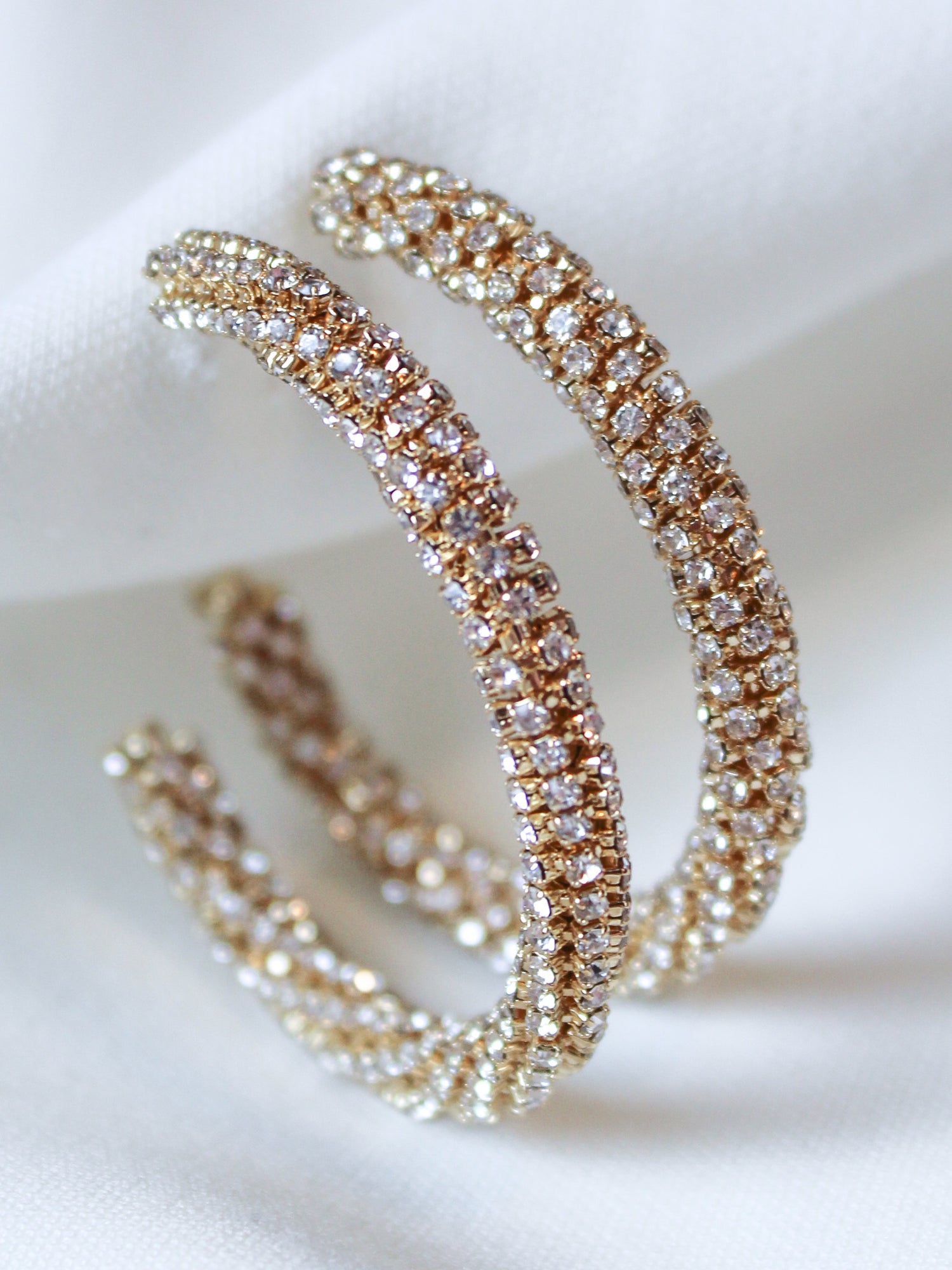 The Riley Hoop earrings by Kinsey Designs feature gold plating and are encrusted with sparkling cubic zirconia, shown on a white fabric background.