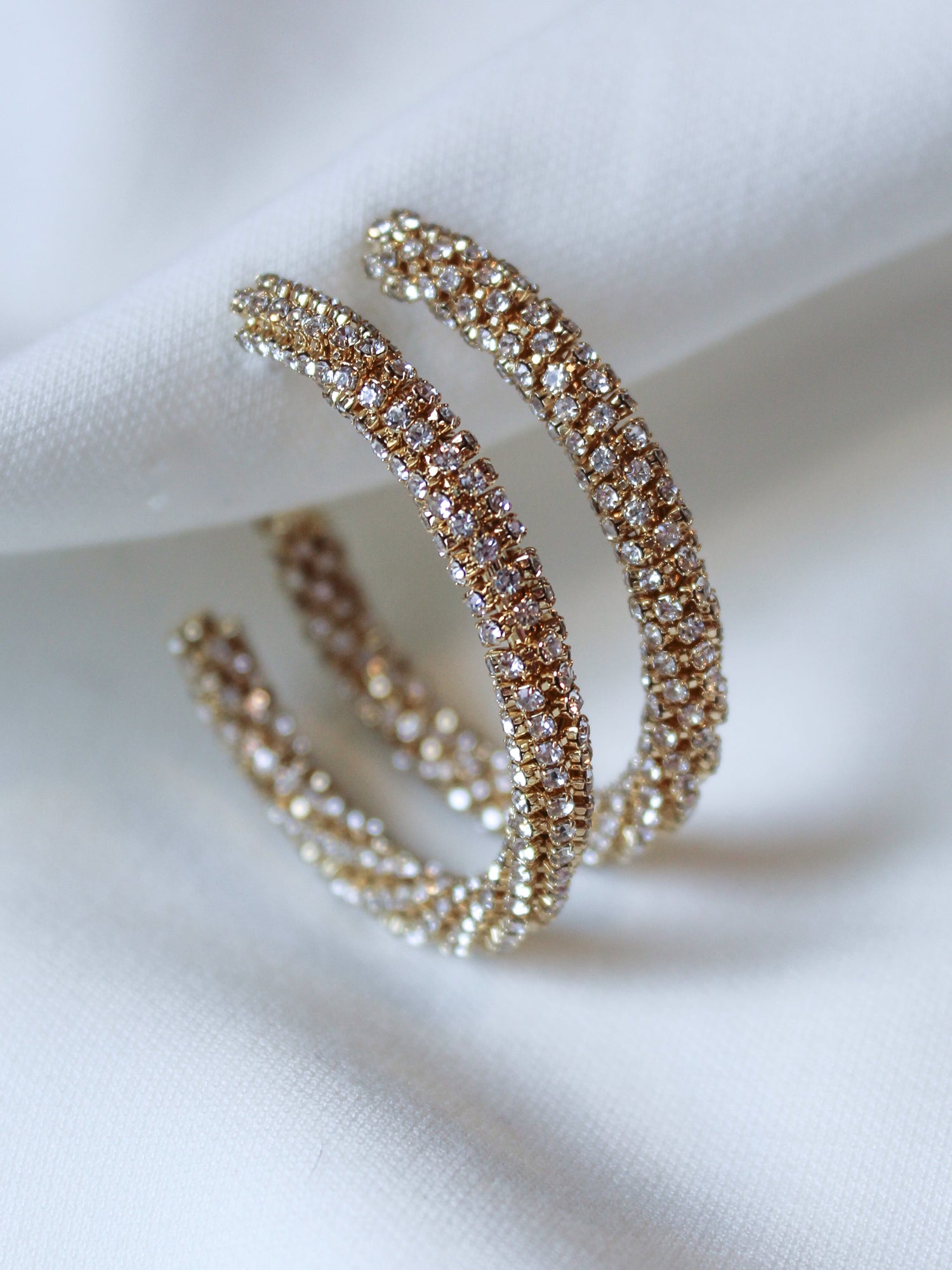 The Riley Hoop by Kinsey Designs features gold-plated, diamond-encrusted double hoops with sparkling cubic zirconia, displayed elegantly on white fabric—an exquisite find at our sample sale.