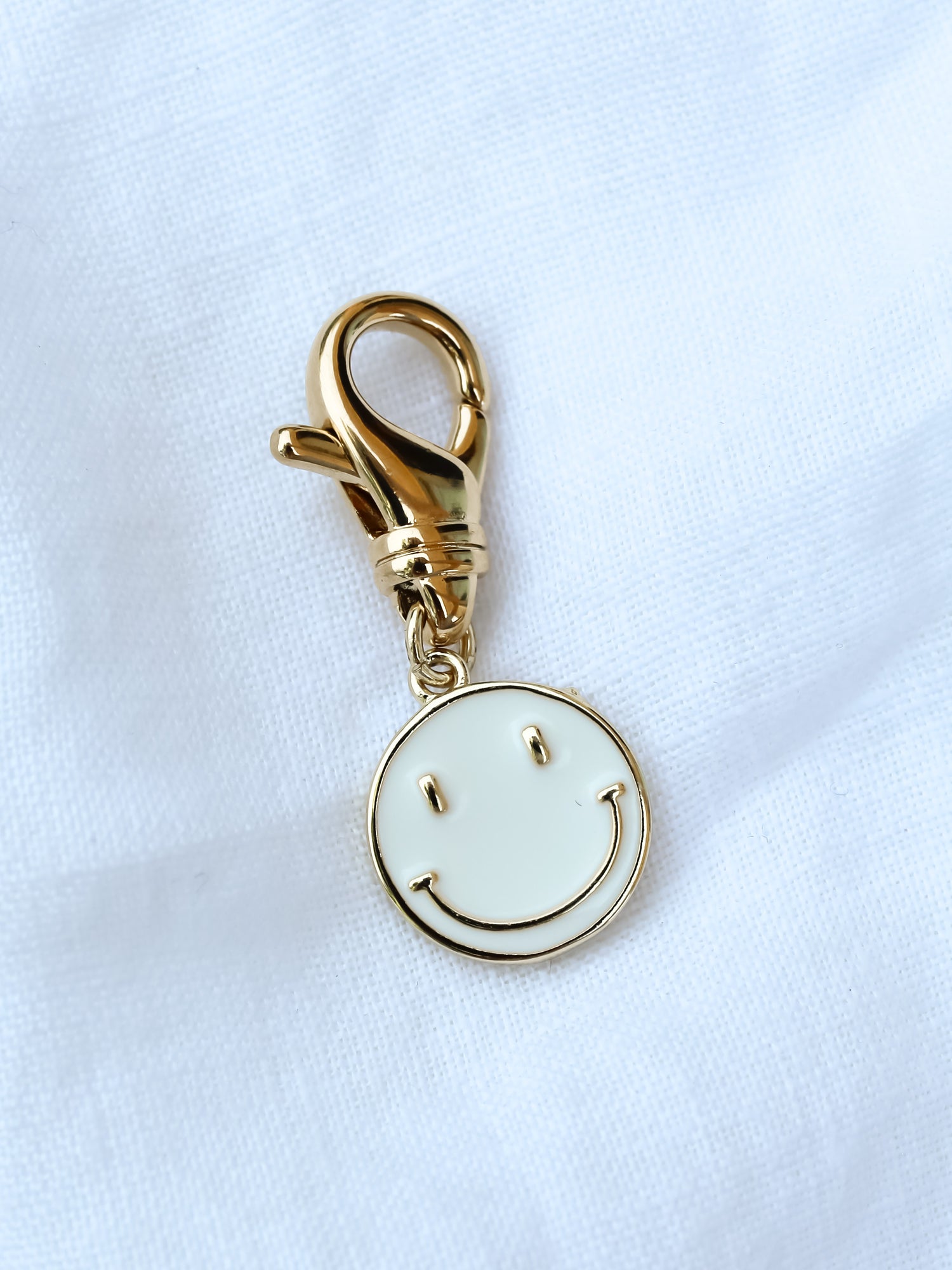 Kinsey Designs' Smiley Shoe Charm features a gold keychain with a white smiley face, shown on a white fabric background. Pair it with our gold-filled earrings and shop now during our exclusive sample sale!.