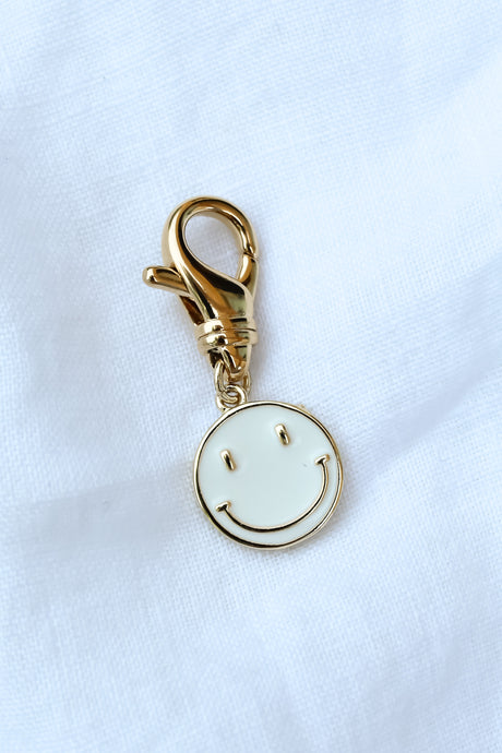 Kinsey Designs' Smiley Shoe Charm features a gold keychain with a white smiley face, shown on a white fabric background. Pair it with our gold-filled earrings and shop now during our exclusive sample sale!.