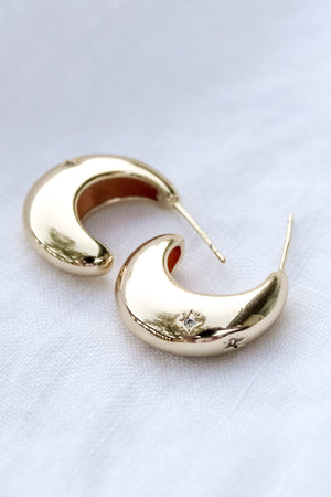 The Swen Earring by Kinsey Designs Archives features a .75" wide crescent shape in 14k gold fill, sparkling with cubic zirconia, shown on a white fabric background.
