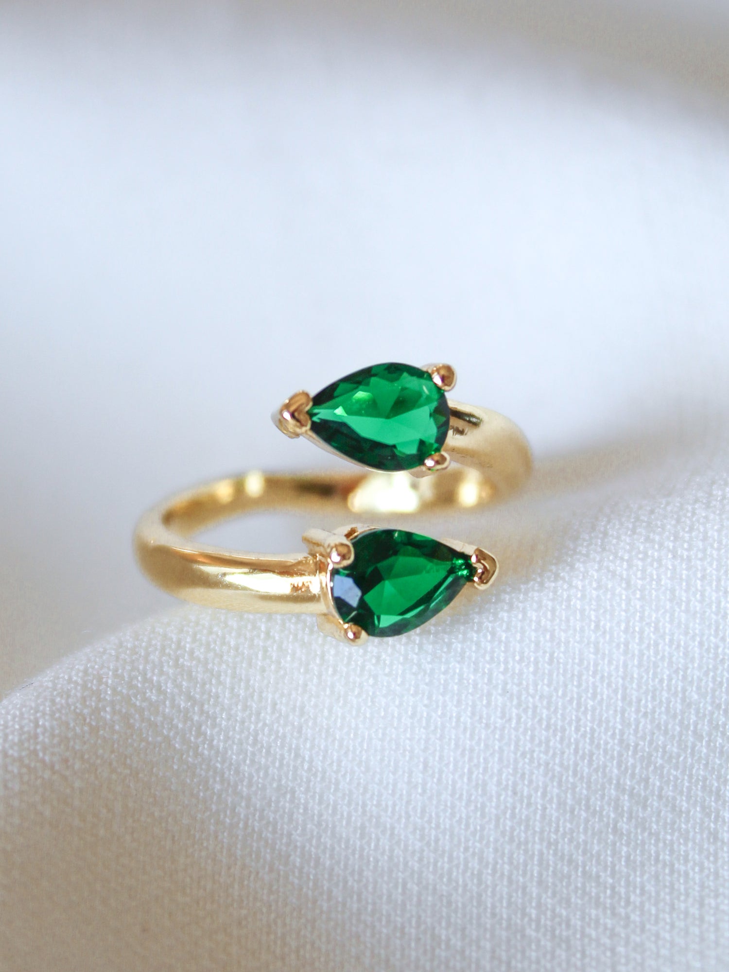 The Teara Ring - Emerald by Kinsey Designs Archives features two pear-shaped green stones and sparkling cubic zirconia accents, elegantly displayed on a white fabric background.