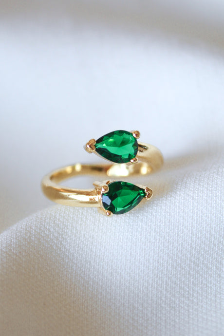 The Teara Ring - Emerald by Kinsey Designs Archives features two pear-shaped green stones and sparkling cubic zirconia accents, elegantly displayed on a white fabric background.