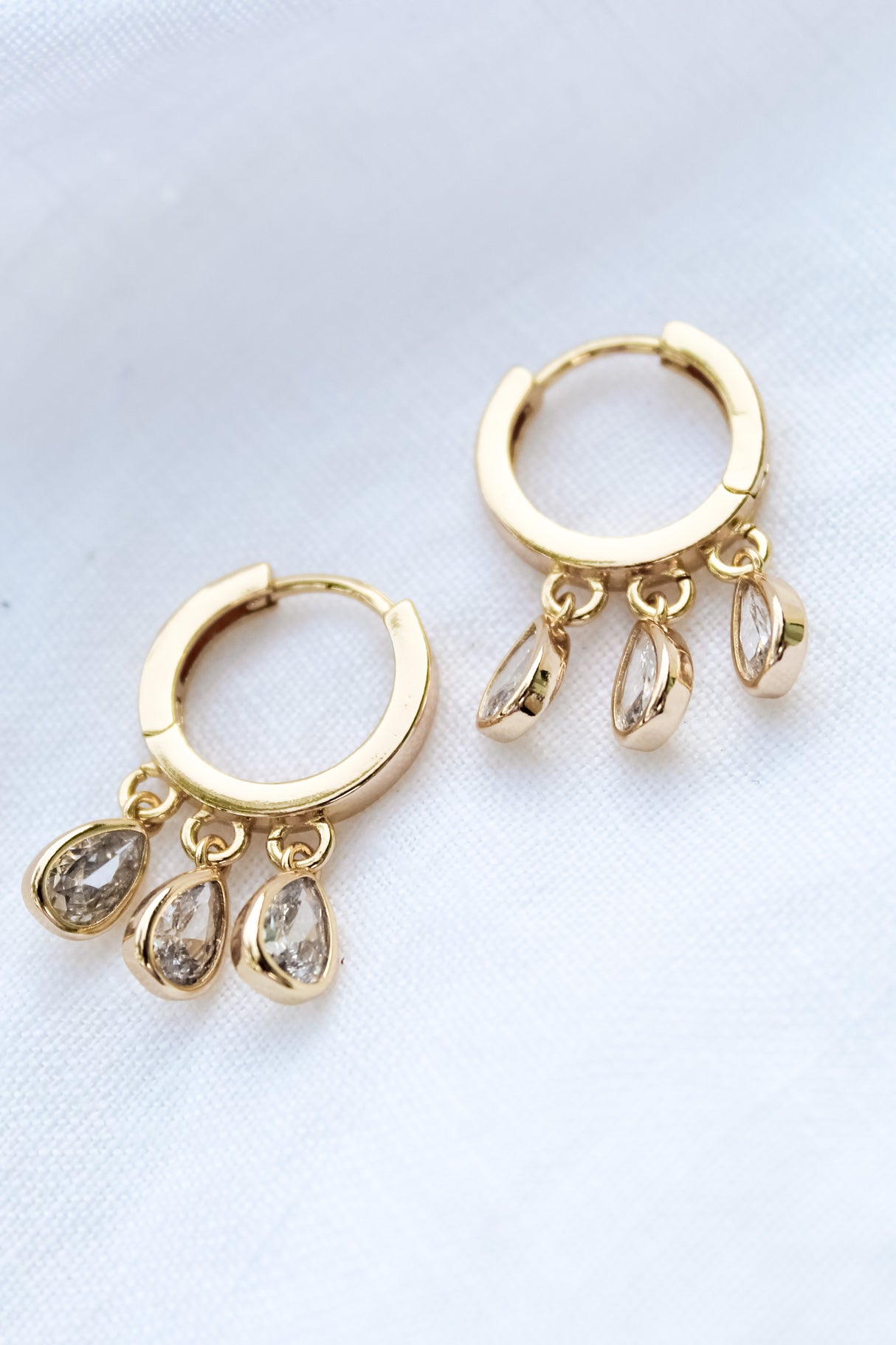 The Tove Huggie by Kinsey Designs Archives features 1-inch gold filled hoops with three teardrop-shaped cubic zirconia charms, shown on white fabric.