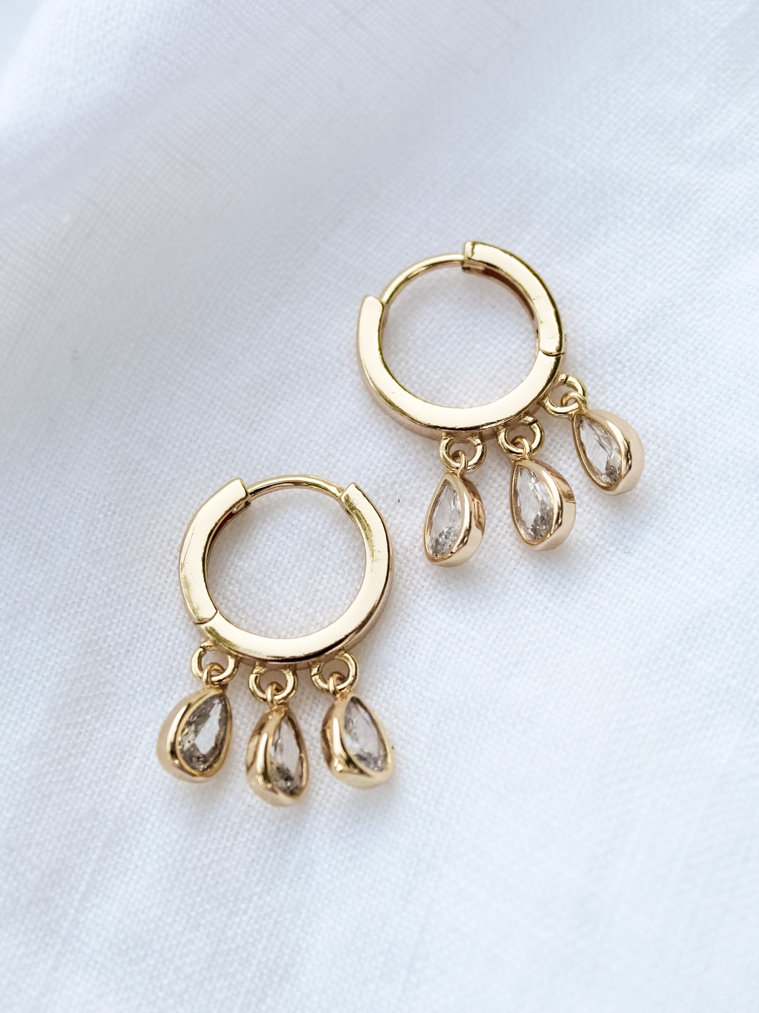 The Tove Huggie by Kinsey Designs Archives are gold-filled hoop earrings, each 1 inch long and featuring three teardrop-shaped cubic zirconia charms, displayed on white fabric.