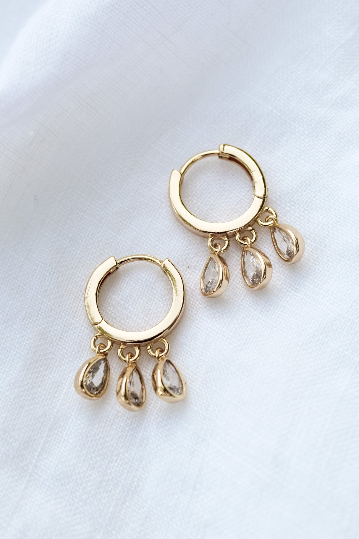 The Tove Huggie by Kinsey Designs Archives are gold-filled hoop earrings, each 1 inch long and featuring three teardrop-shaped cubic zirconia charms, displayed on white fabric.