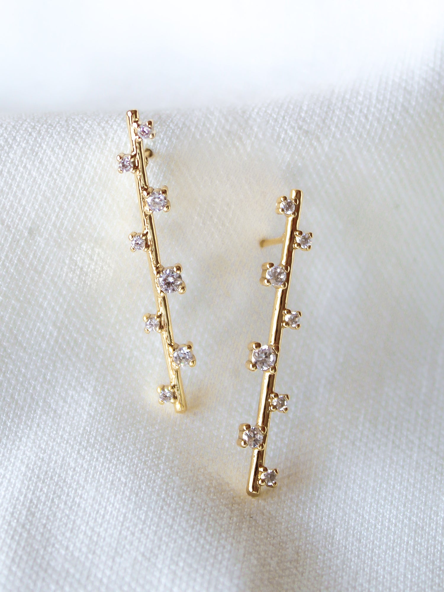 The Walker Earring by Kinsey Designs Archives features 1.5-inch gold-filled drops with vertical rows of small cubic zirconia, shown on a white fabric background. These elegant earrings bring subtle sparkle to any look.