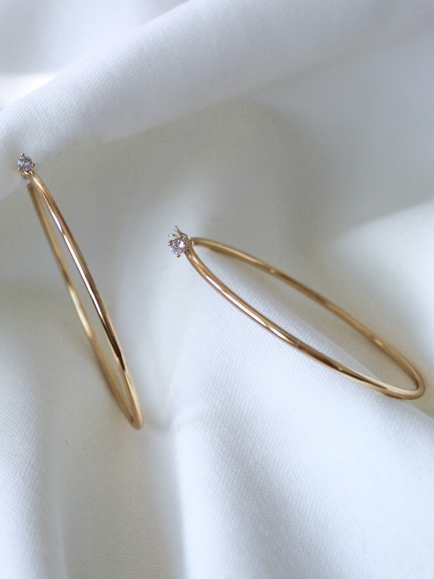 The Xandra Hoop earrings by Kinsey Designs Archives, featuring sparkling cubic zirconia, rest on soft white fabric.