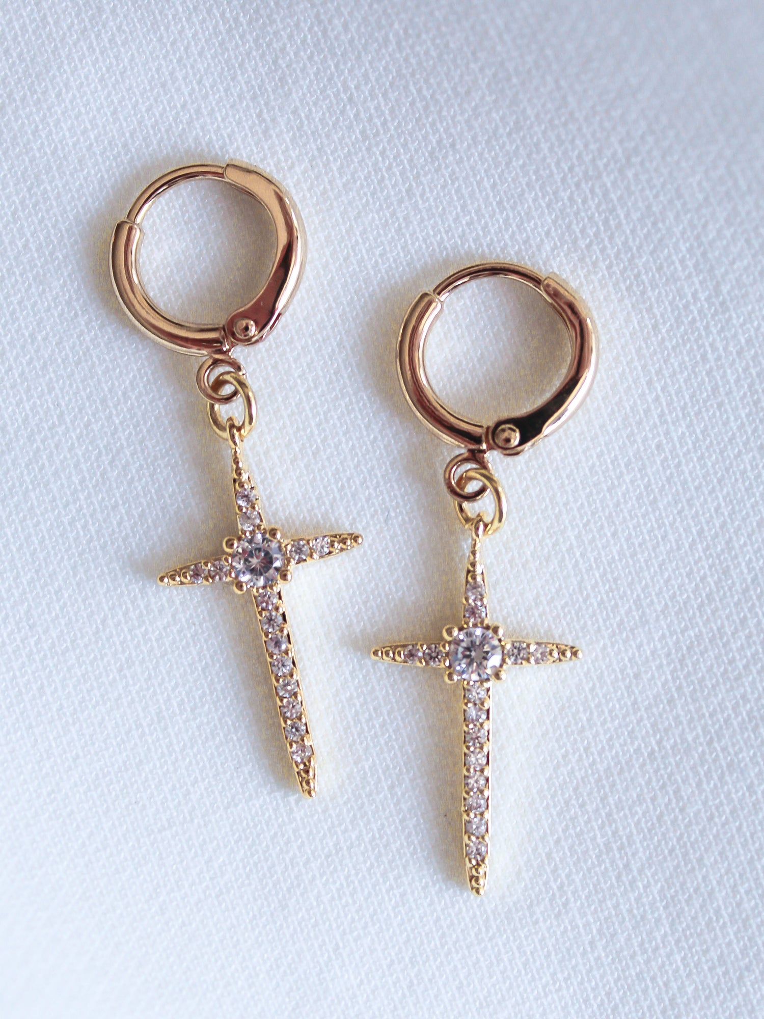 The Zenni Huggie by Kinsey Designs Archives are gold hoop earrings with dangling, jeweled cross pendants featuring sparkling cubic zirconia, displayed on a white textured background.