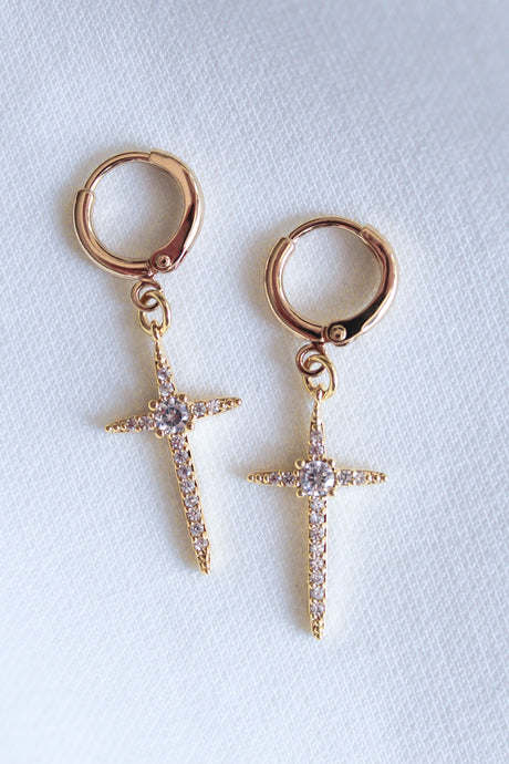 The Zenni Huggie by Kinsey Designs Archives are gold hoop earrings with dangling, jeweled cross pendants featuring sparkling cubic zirconia, displayed on a white textured background.
