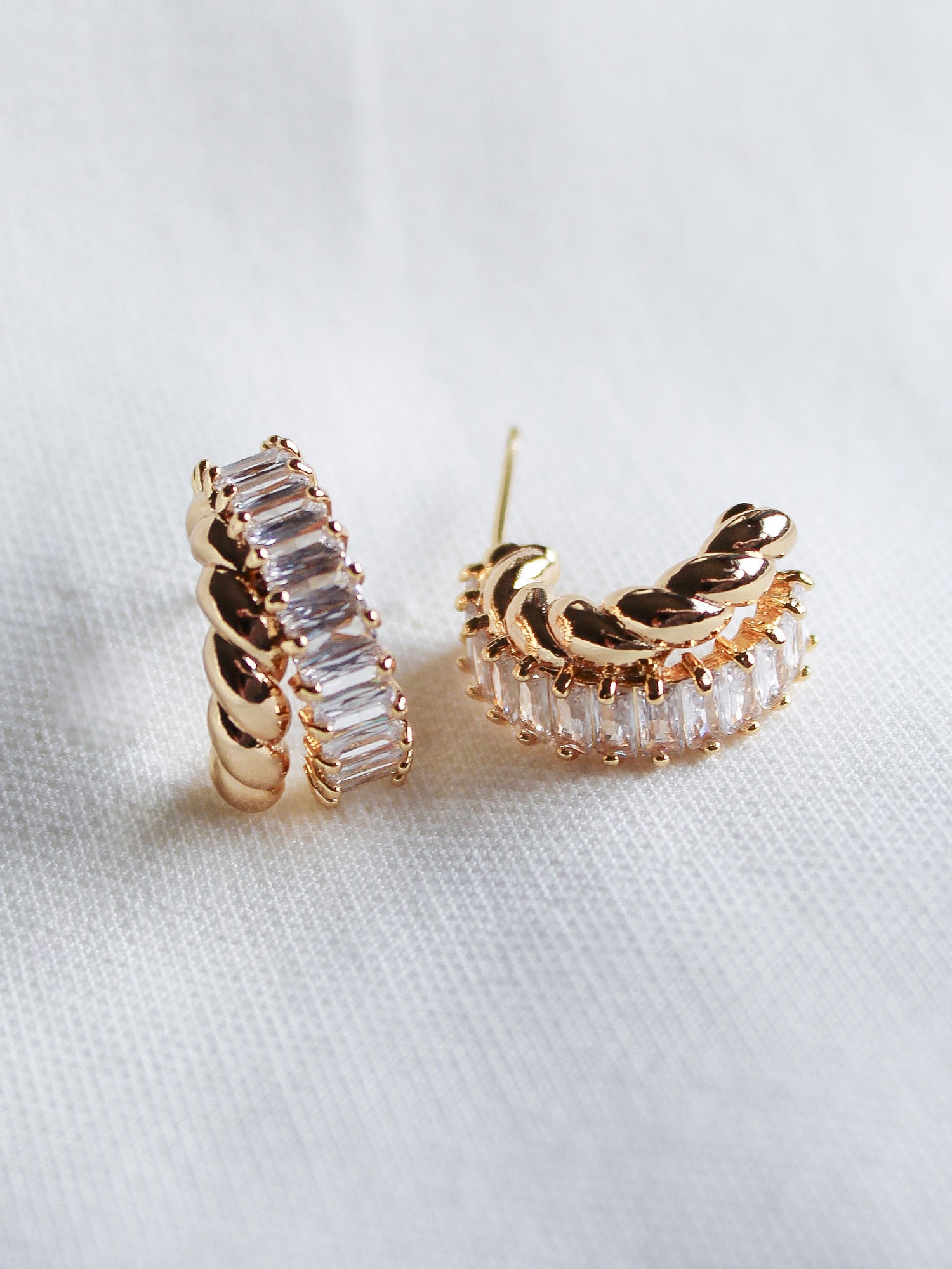 The Zuri Hoop by Kinsey Designs features gold-filled hoops with clear rectangular cubic zirconia in a twisted design, displayed on white fabric.